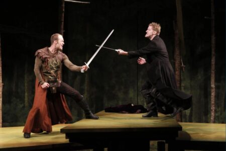 Two actors in medieval costumes perform a dramatic sword fight on stage, one in brown and the other in black, set against a backdrop of trees and a wooden platform.