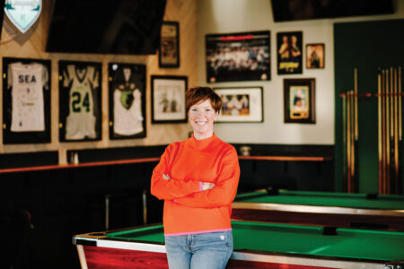 A person with short hair wearing a bright orange sweater and jeans stands smiling with arms crossed in front of pool tables, surrounded by framed jerseys and sports memorabilia on the walls.