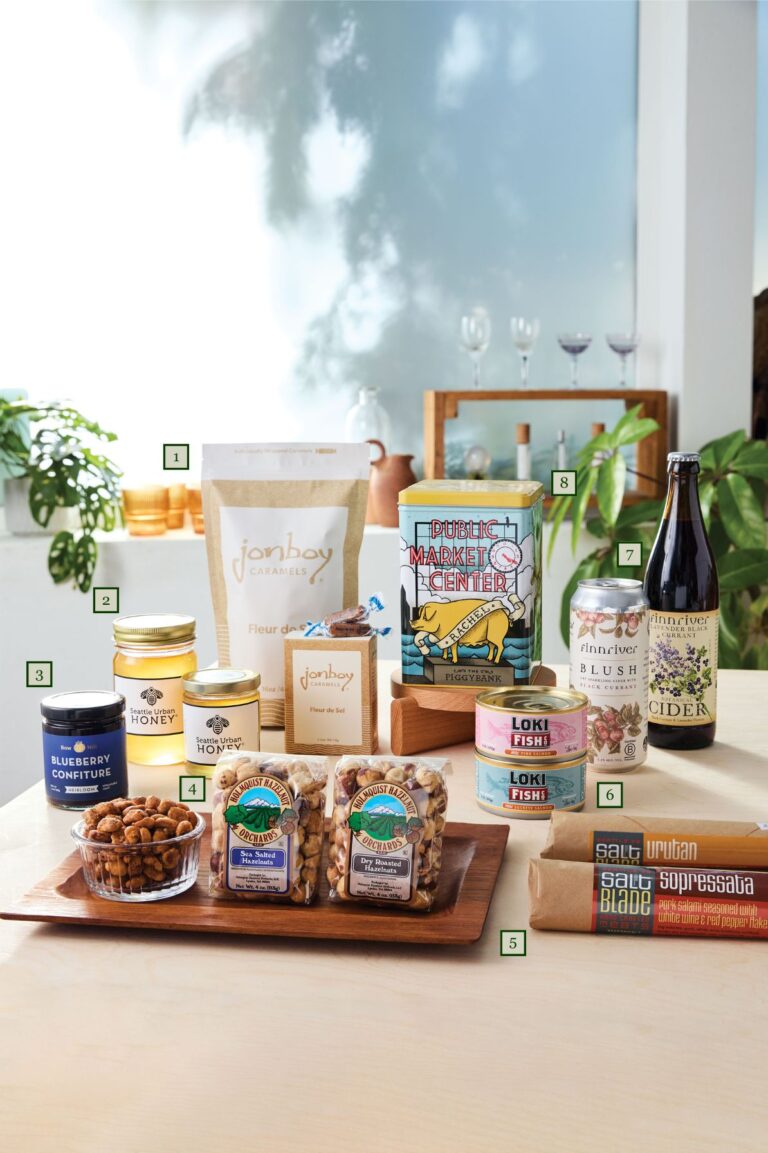 A display of gourmet food items on a light wooden table, including jars, tins, packages of snacks, almond nuts, a bottle of cider, and cheese, with potted plants and wine glasses in the background. Each item is numbered.