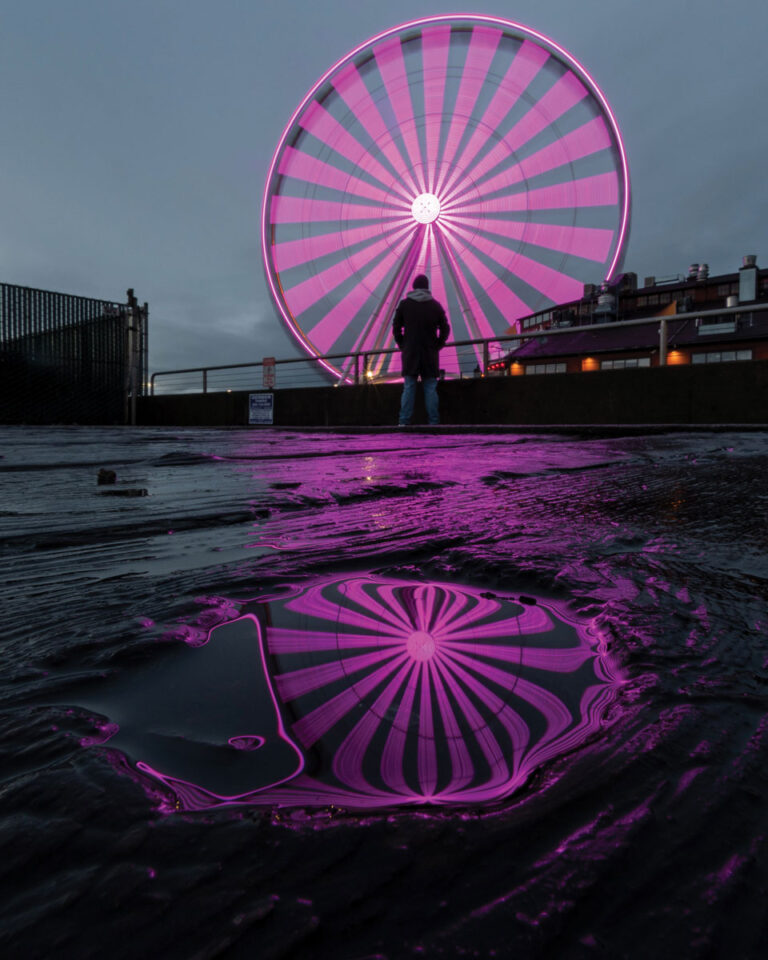A brightly lit pink ferris wheel is reflected in a large puddle on the ground at dusk. A person stands in front of the ferris wheel, with buildings and a cloudy sky in the background.