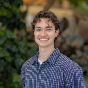 A young man with wavy brown hair, wearing a blue plaid shirt over a white t-shirt, smiles at the camera. The background is blurred greenery, suggesting an outdoor or garden setting.