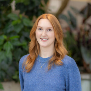 A young woman with long red hair, wearing a blue sweater and a necklace, smiles while standing in front of green plants.