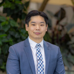 A man in a blue suit, white shirt, and patterned tie sits in front of leafy green plants, looking at the camera with a neutral expression.