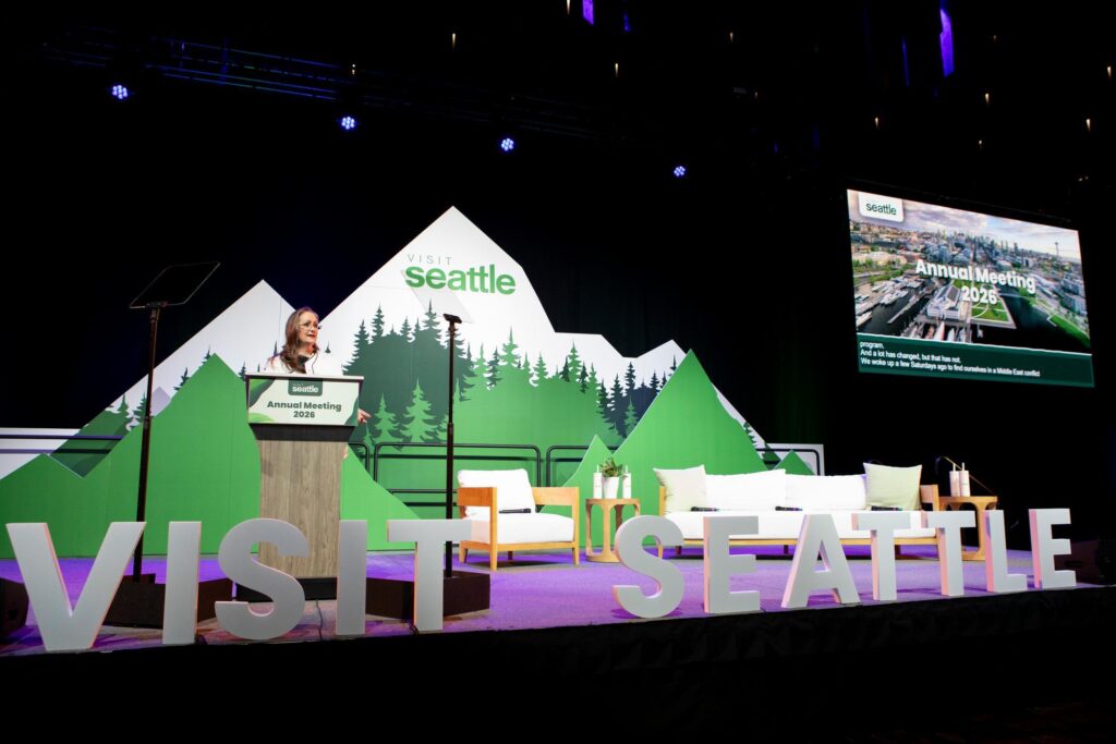 A woman stands at a podium on stage at the "Visit Seattle" Annual Meeting 2023. The stage is decorated with mountain graphics, large text reading "VISIT SEATTLE," and a screen displaying meeting information.