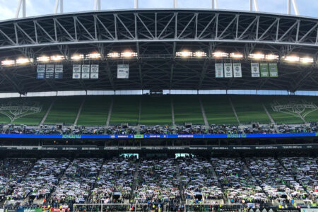 A soccer match is in progress at a large stadium with fans filling the stands, many holding scarves. The stadium roof arches overhead, and sunlight illuminates the field and audience.