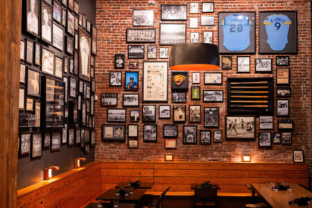 A cozy restaurant with brick walls covered in framed photos, sports memorabilia, and two blue baseball jerseys. Wooden benches and tables are arranged below warm lighting, creating an inviting atmosphere.