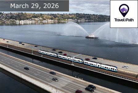 A light rail train crosses the floating bridge over water with cars on adjacent lanes; a boat sprays water in the background. The date "March 29, 2026" and a "Travel the Path" logo are displayed at the top.