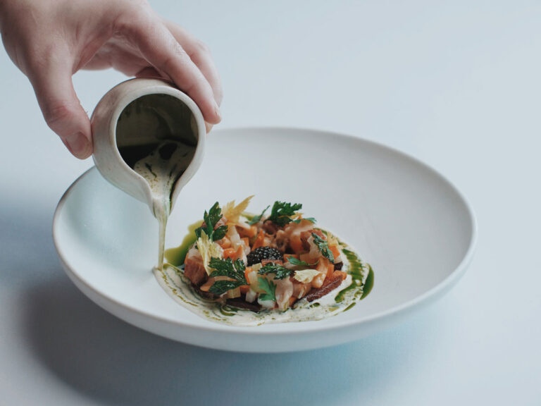 A hand pours green sauce from a small jug onto an artfully plated dish with herbs, caviar, and garnishes on a white plate against a light background.