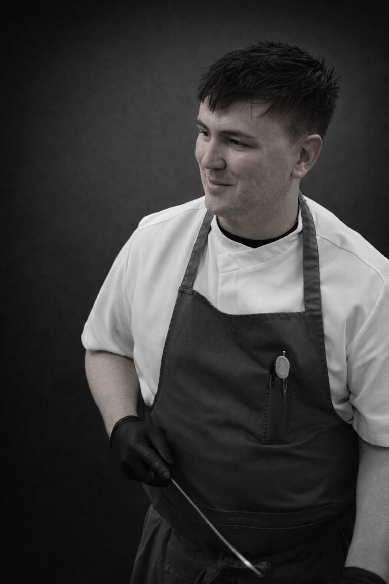 A young chef wearing a white shirt, dark apron, and black gloves holds a kitchen utensil, smiling slightly while looking to the side against a dark background.