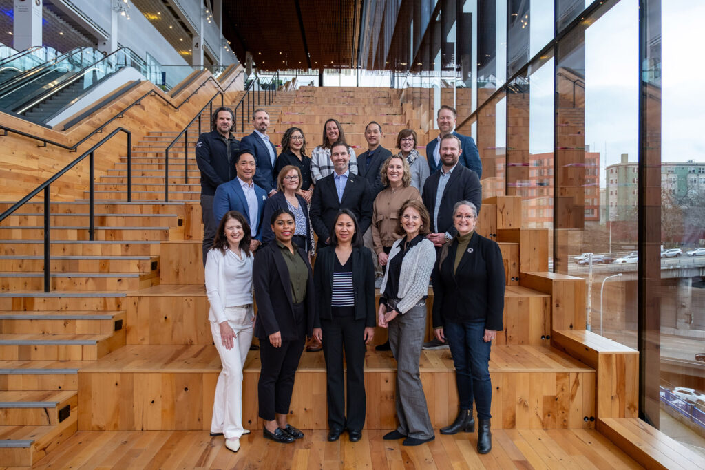 A group of 19 professionally dressed people stand and smile on large wooden steps inside a modern building with tall glass windows and an urban view outside.