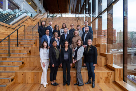 A group of 19 professionally dressed people stand and smile on large wooden steps inside a modern building with tall glass windows and an urban view outside.