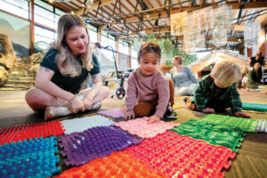 A woman and two young children sit on the floor, exploring colorful textured sensory mats in a bright, spacious indoor play area with natural light and wooden beams.