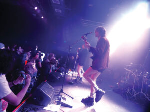 A band performs on a brightly lit stage in front of a cheering crowd. The guitarist and vocalist are in the foreground, while fans take photos and enjoy the live music in the dimly lit venue.