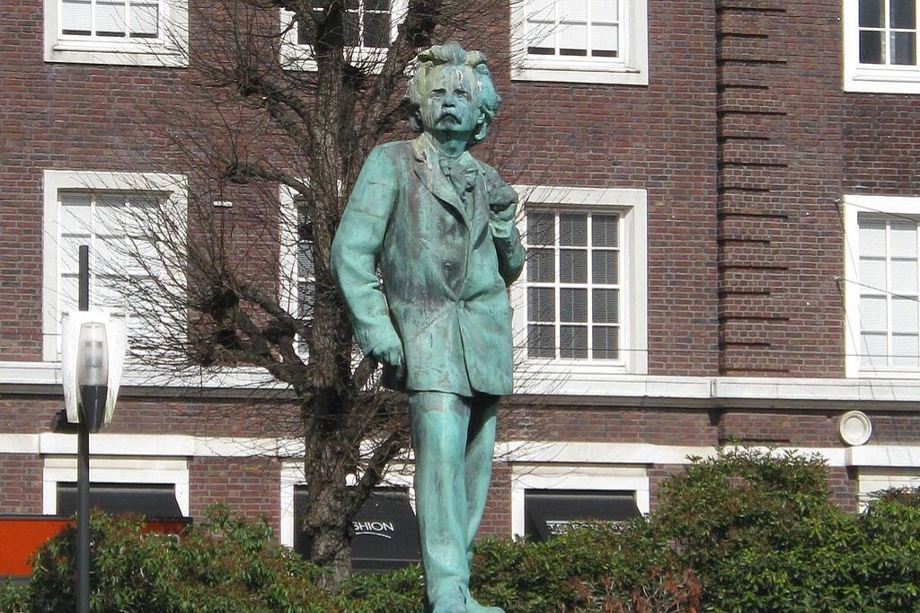 A bronze statue of a man with wavy hair and a mustache stands in front of a brick building with white window frames. Leafless branches are visible behind the statue. Shrubs grow at the base.