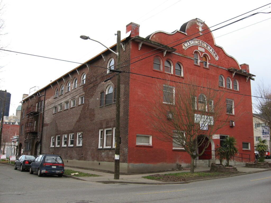 A three-story brick building with arched windows and a sign reading "Washington Hall" above the entrance, located on a street corner with parked cars and leafless trees nearby.