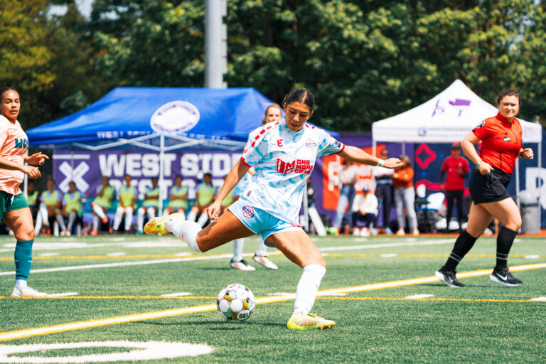 A soccer player in a white and light blue uniform kicks a ball on the field during a Seattle sports match, with other players, referees nearby, and tents set up along the sideline in the background.