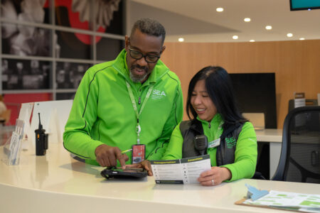 Two airport staff members in green uniforms stand behind an information desk, smiling and looking at a tablet and a brochure together. The background includes informational materials and a computer monitor.